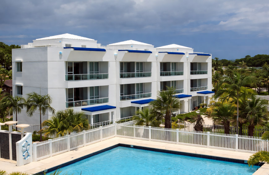 Modern three-story condominium building with blue awnings, private balconies, and resort-style pool in a tropical coastal setting.