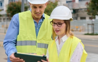 Engineers in hard hats and yellow safety vests look at a tablet.